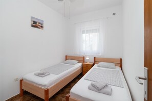 A small, clean room with two single beds, a bedside table with a lamp, a window with sheer curtains, and a small painting on the wall. - Apartments Ivica (Vir)