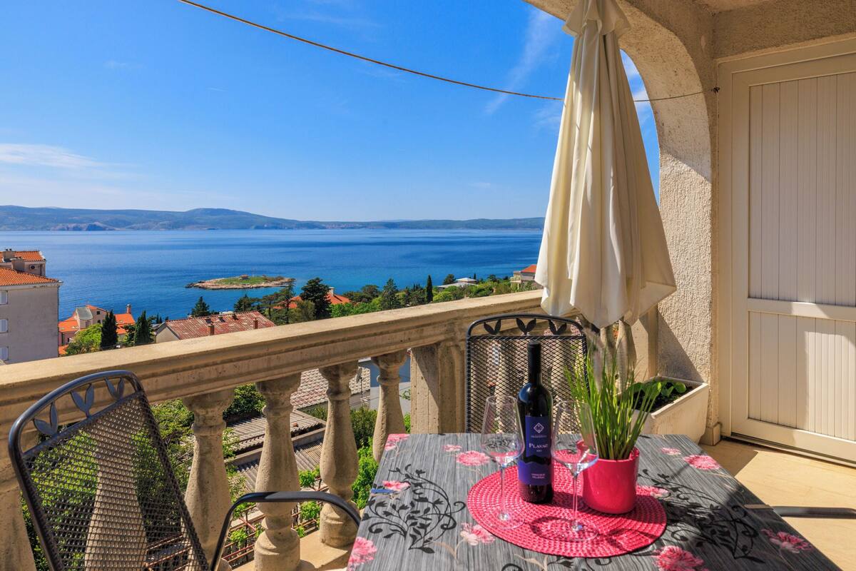 Un balcon avec une table, des chaises, un pot de fleurs et une bouteille de vin.