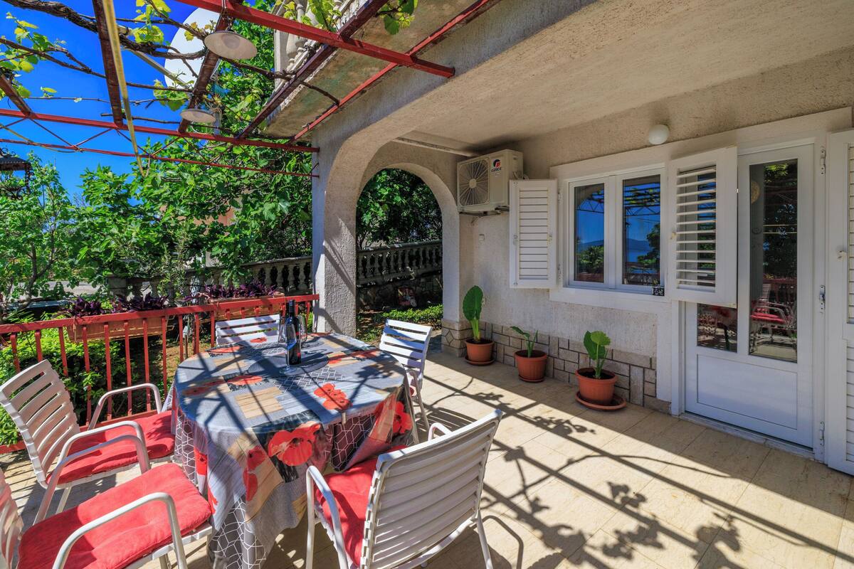 Une terrasse extérieure avec une table dressée pour le repas, entourée de chaises blanches, de plantes en pot et d’une rambarde rouge.