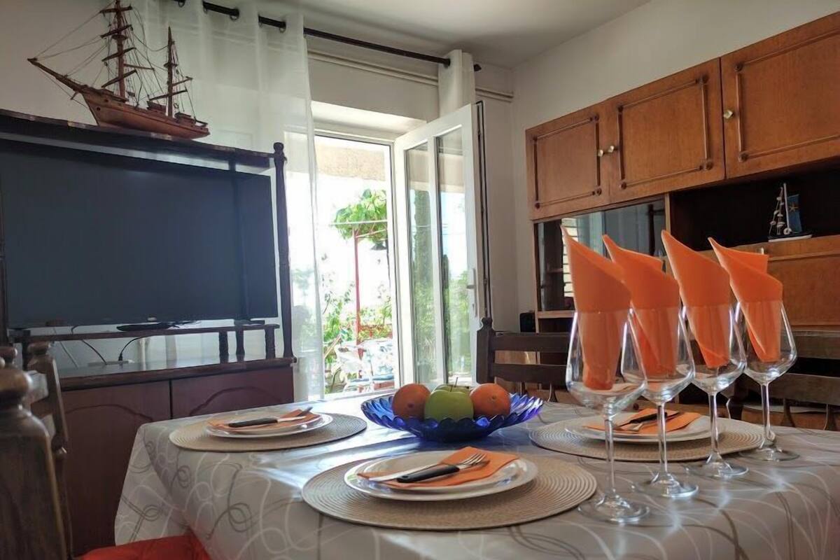 Un coin repas avec une table dressée pour quatre personnes, une maquette de bateau accrochée au mur et une vue sur l’extérieur.