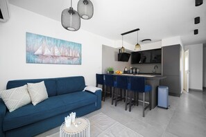 A modern living room with a blue sofa, a dining area with blue chairs, a kitchen with a black countertop, and a painting of sailboats on the wall. - House Luxury Konforta (Sibenik)