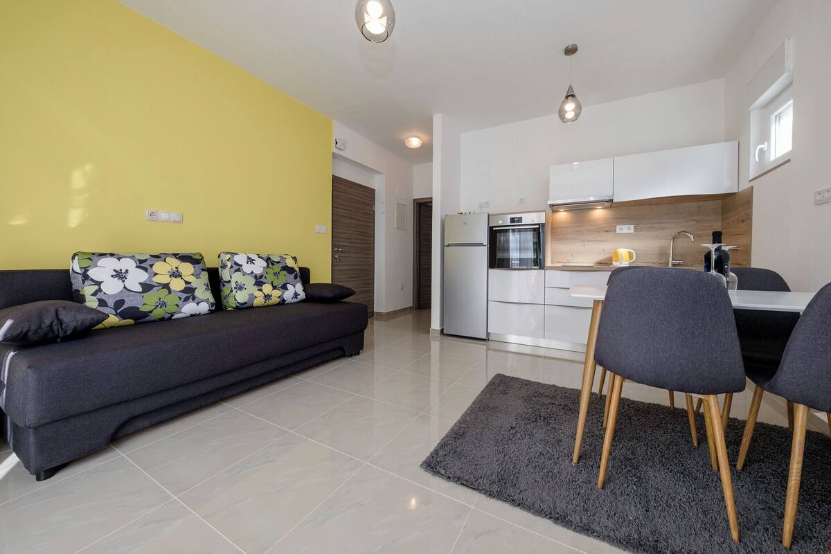 A modern living room with a sofa and chairs, a kitchen with white cabinets, and a grey rug.