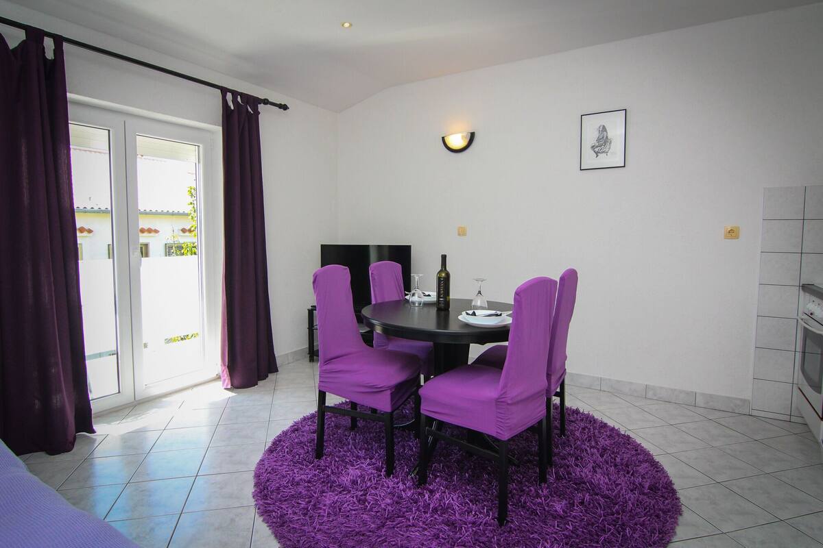 A dining area with a round table, purple chairs, and a purple rug. There is a TV on a stand, a wine bottle, and a glass on the table.