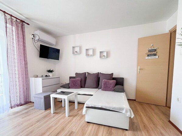 Quarto moderno e bem iluminado com sofá, uma pequena mesa, TV fixada na parede e uma porta com a inscrição 'Cozinha Onde Amigos São Bem-Vindos'.