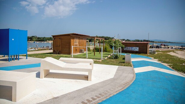 Eine Küstenlandschaft mit einem Holzhaus, einer blauen Konstruktion und einer weißen Bank.