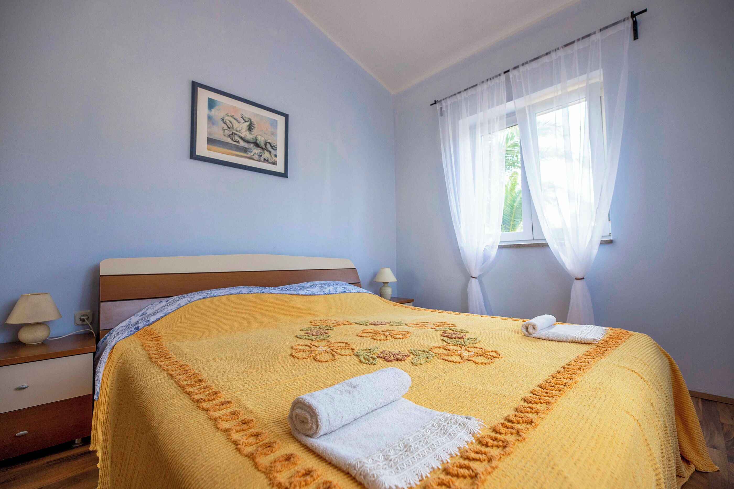 A neatly made bed with a floral-patterned bedspread, a bedside table with a lamp, and a window with sheer curtains.