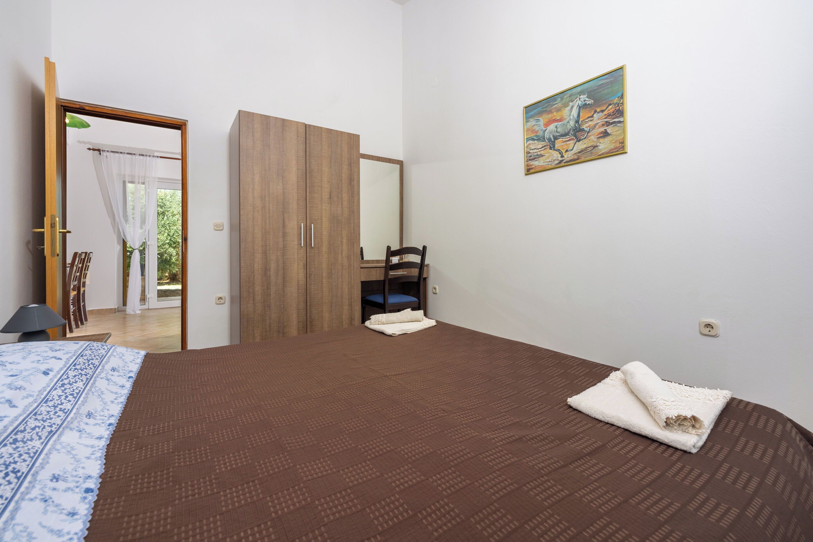Un dormitorio con colcha marrón, un armario de madera, una silla y un cuadro en la pared.