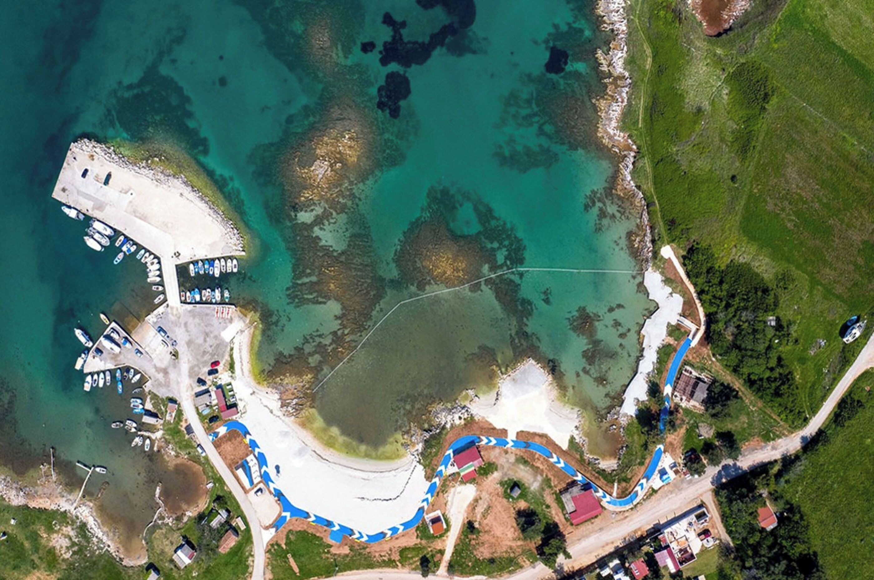 Una vista aerea di una zona costiera con un porto turistico, una spiaggia sabbiosa e una strada tortuosa.