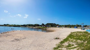Ein Strand mit blauem Himmel, felsiger Küste und einigen Gebäuden im Hintergrund.