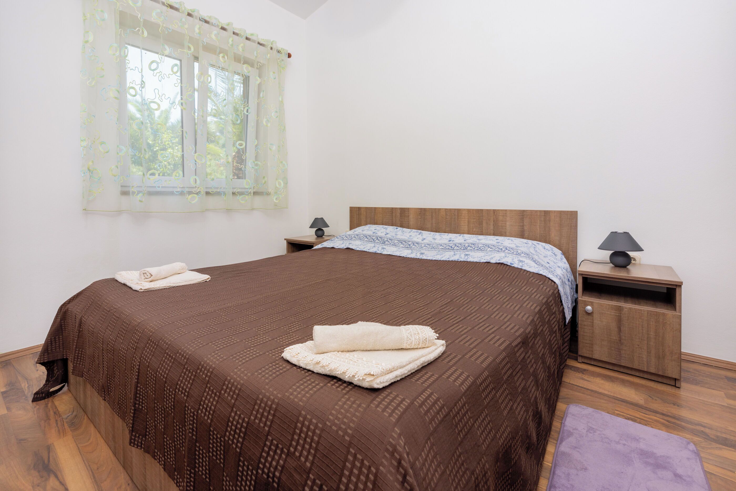 Ein ordentlich bezogenes Bett mit einer braunen, gemusterten Tagesdecke und einem weißen, darübergelegten Handtuch. Am Bett befindet sich ein Nachttisch mit einer Lampe.