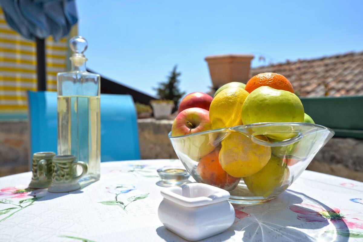 A table with a bowl of fruit, a salt shaker, and a small dish, set outdoors with a clear sky and a building in the background.