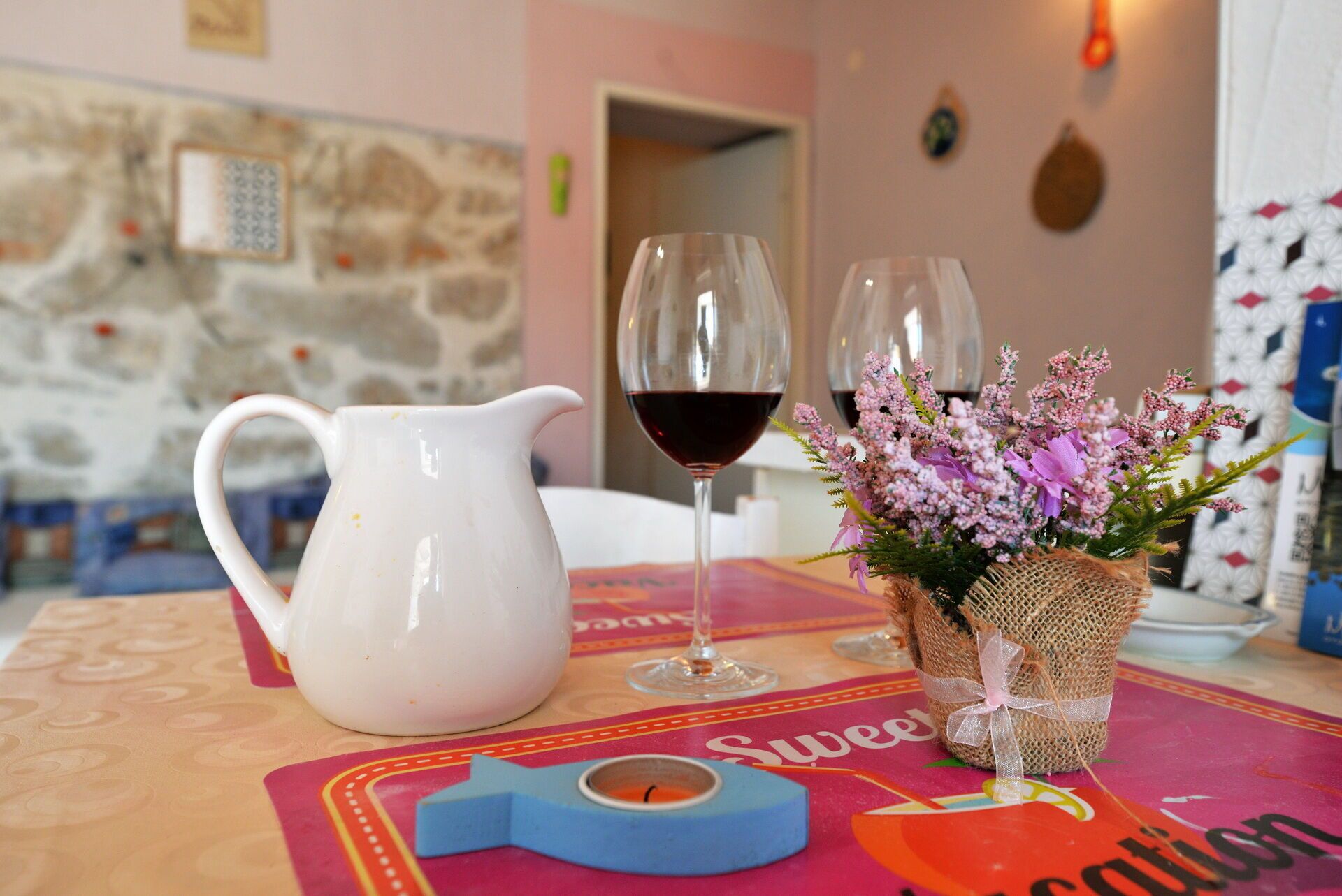 A table with a white pitcher, two wine glasses with red wine, a small vase with pink flowers, and a colorful placemat with the word "Sweet."