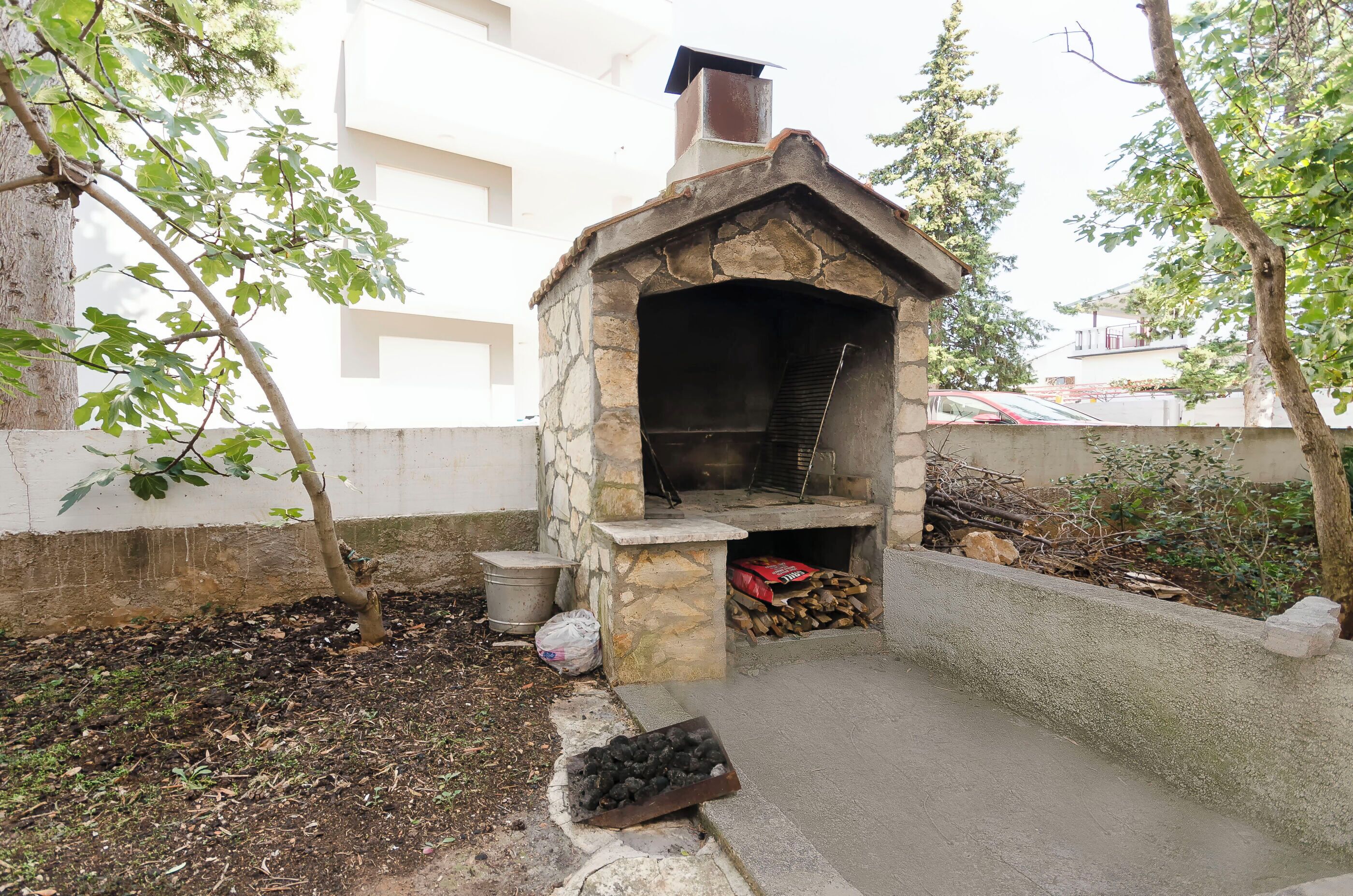 A stone-built outdoor oven with a chimney, surrounded by trees and a concrete wall.