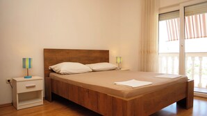 A bedroom with a wooden bed, two bedside tables, a nightstand, a window with curtains, and a view of greenery outside. - Apartments Vlade (Primosten)