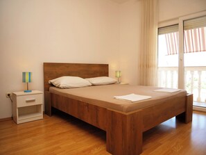 A bedroom with a wooden bed, two bedside tables, a nightstand, a window with curtains, and a view of greenery outside. - Apartments Vlade (Primosten)