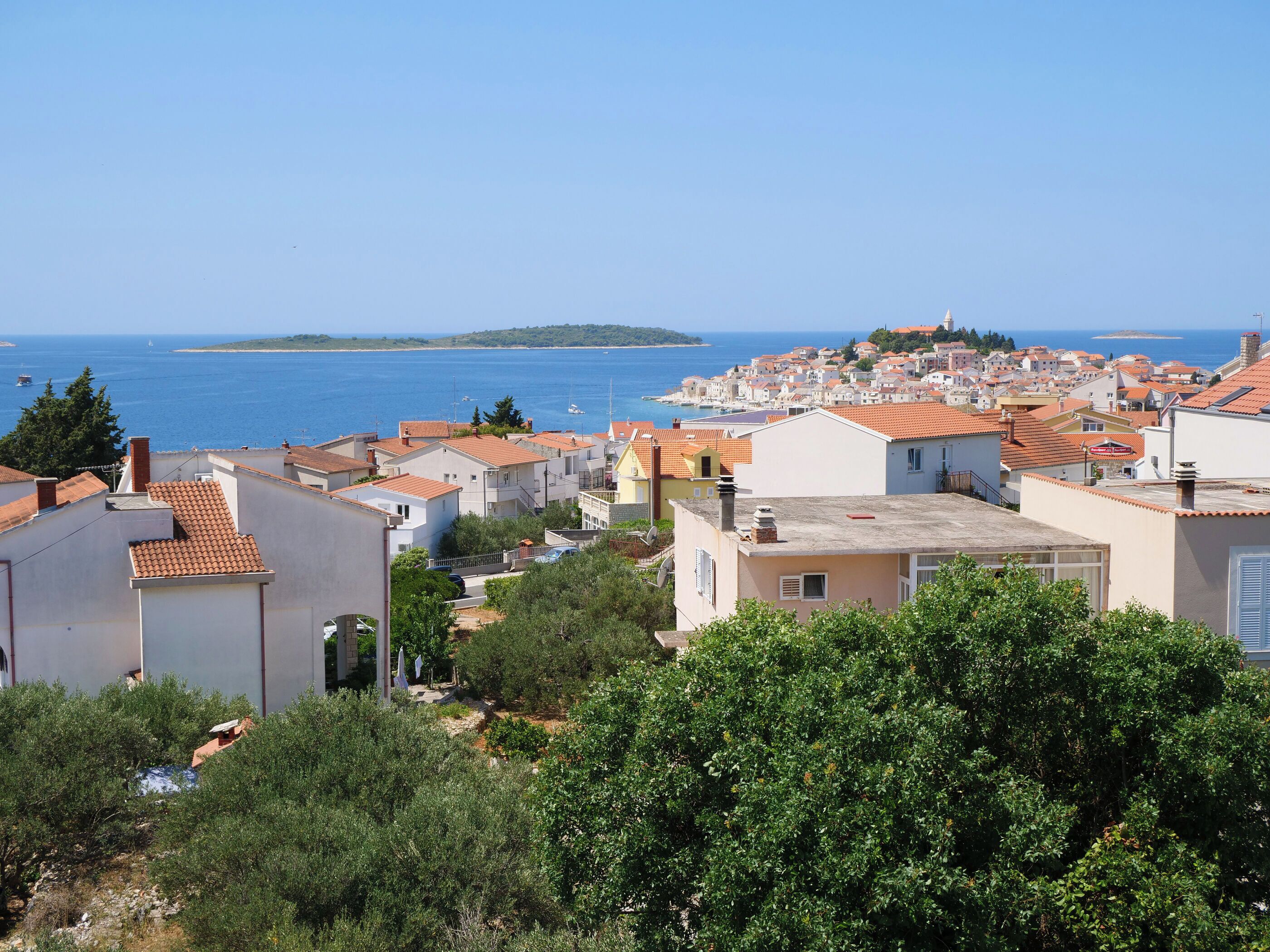 A coastal town with terracotta-roofed buildings, a clear blue sea, and a distant island.