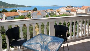 A balcony with a glass table and two chairs, overlooking a coastal town with houses and a sea view. - Apartments Vlade (Primosten)