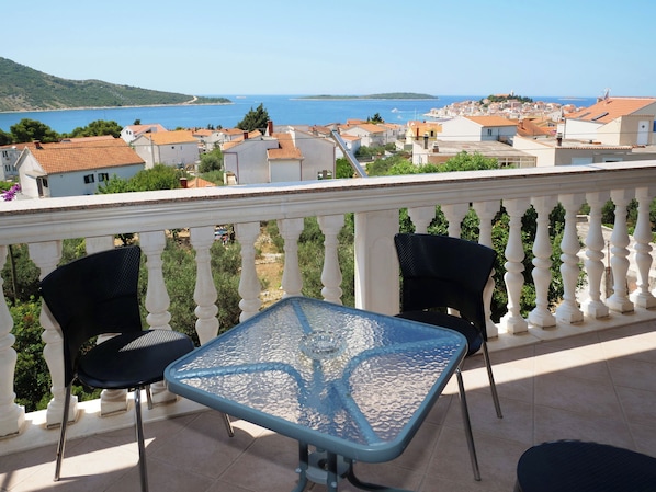 A balcony with a glass table and two chairs, overlooking a coastal town with houses and a sea view. - Apartments Vlade (Primosten)