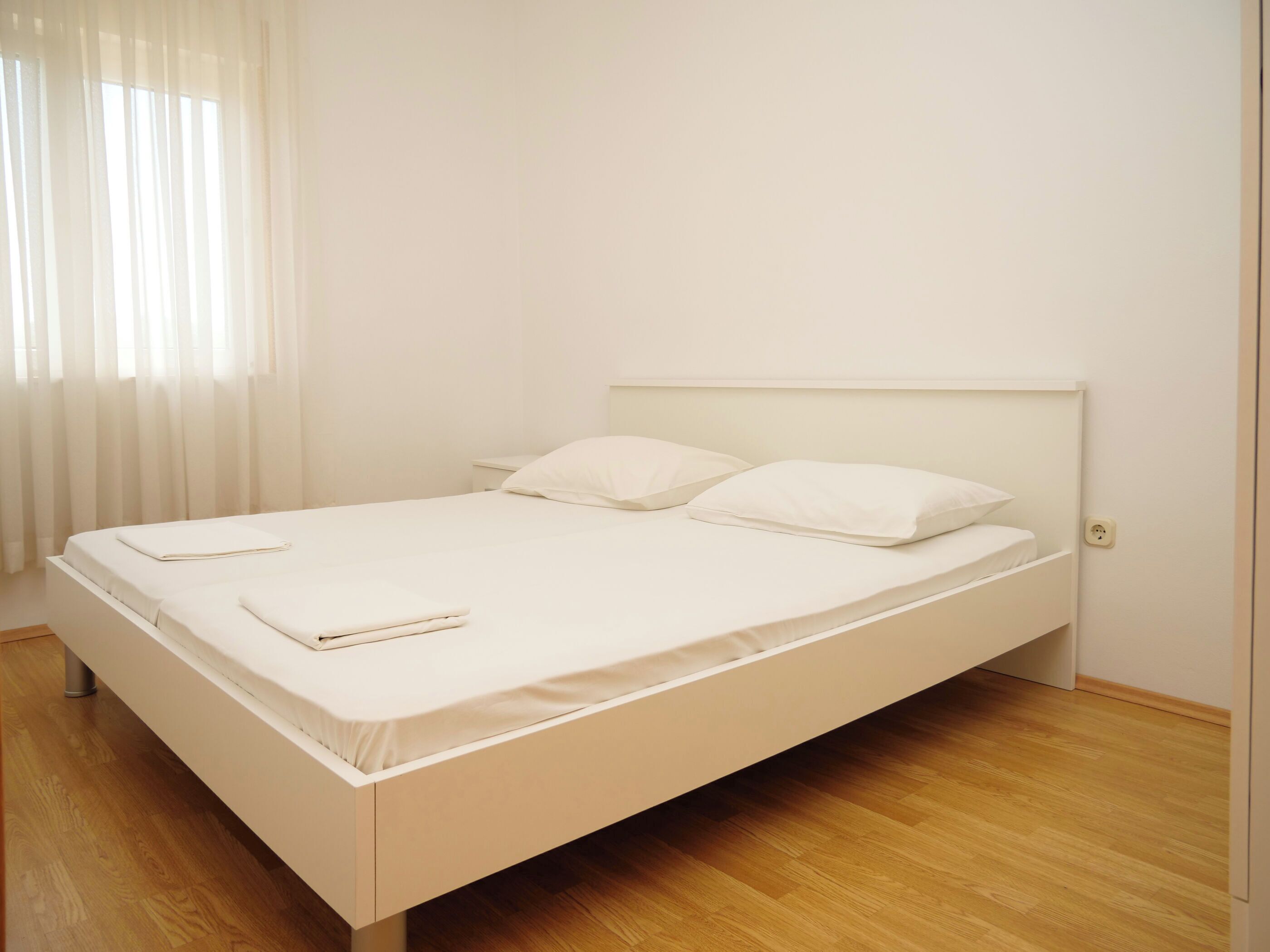 A single-bed room with a wooden floor, white bedding, and a window with sheer curtains.
