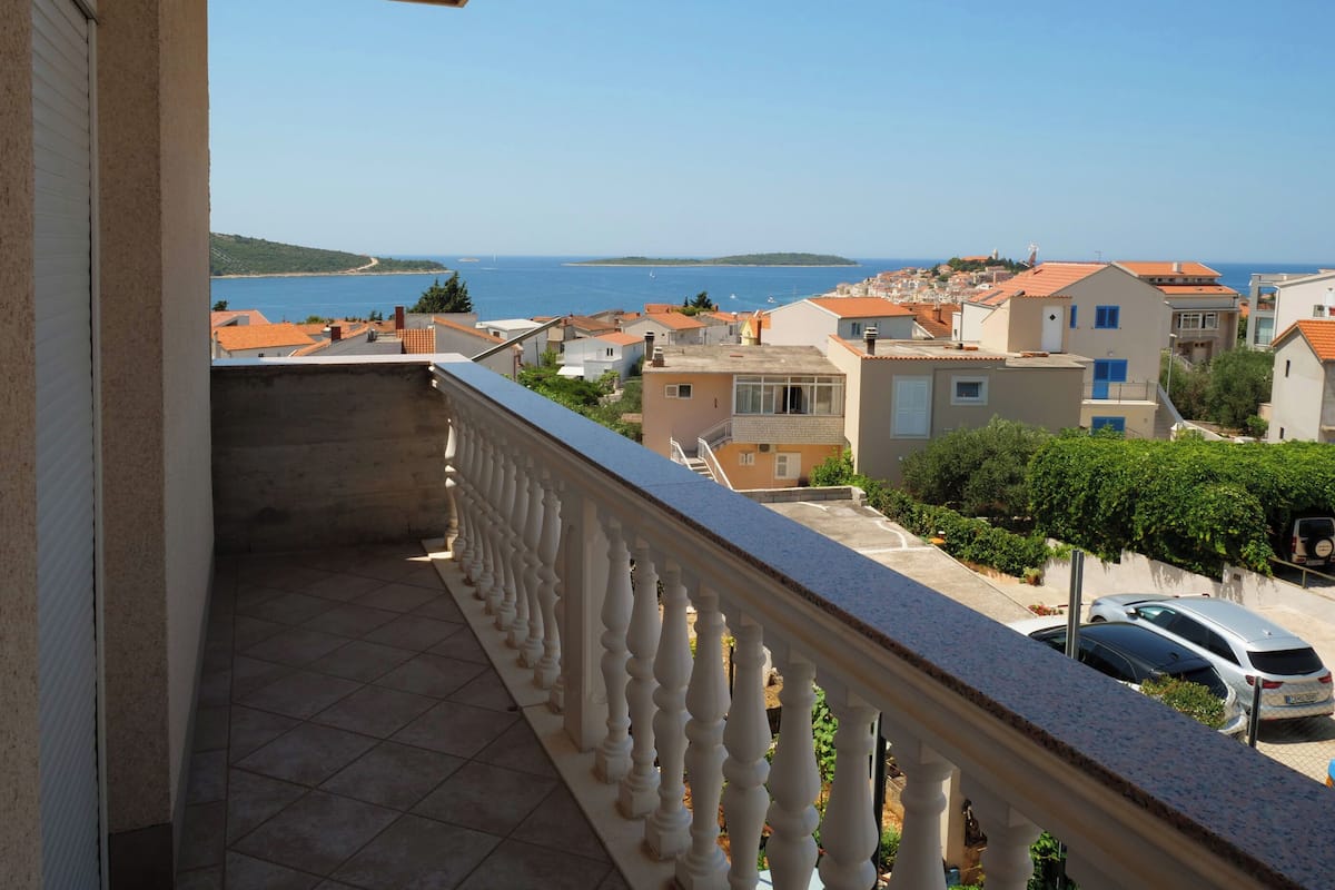 A balcony with a tiled floor and white balustrade overlooking a coastal town with red-roofed buildings and a clear blue sea.