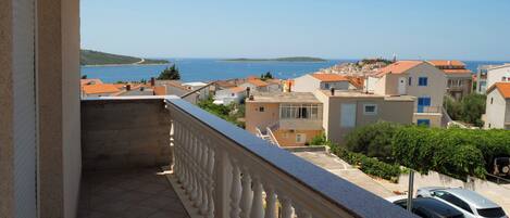 A balcony with a tiled floor and white balustrade overlooking a coastal town with red-roofed buildings and a clear blue sea.