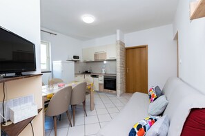 A modern living room with a flat-screen TV, a dining area with a table and chairs, and a kitchen with a stove and cabinets. - Apartments Miranda (Lopar)