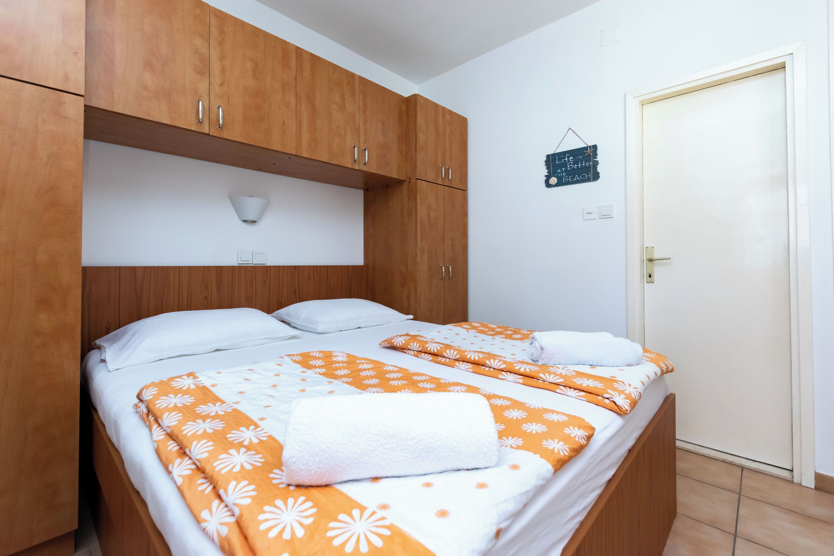 Una cama doble con ropa de cama blanca y anaranjada con motivos florales, cabecera de madera y una pared blanca con un letrero pequeño.