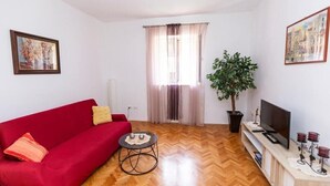 Ein Wohnzimmer mit einem roten Sofa, einem kleinen runden Tisch, einer Zimmerpflanze, einem Fernseher auf einem Schrank und einem Fenster mit Vorhängen.