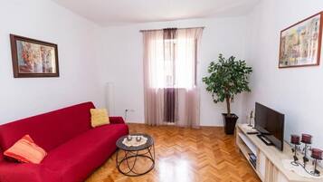Sala de estar com sofá vermelho, uma mesinha redonda, planta em vaso, televisão em um móvel e janela com cortinas.