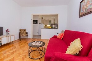Un salon avec un canapé rouge, une petite table basse ronde et un téléviseur posé sur un meuble. On y voit une chaise à motifs floraux et, par une porte ouverte, on aperçoit la cuisine.