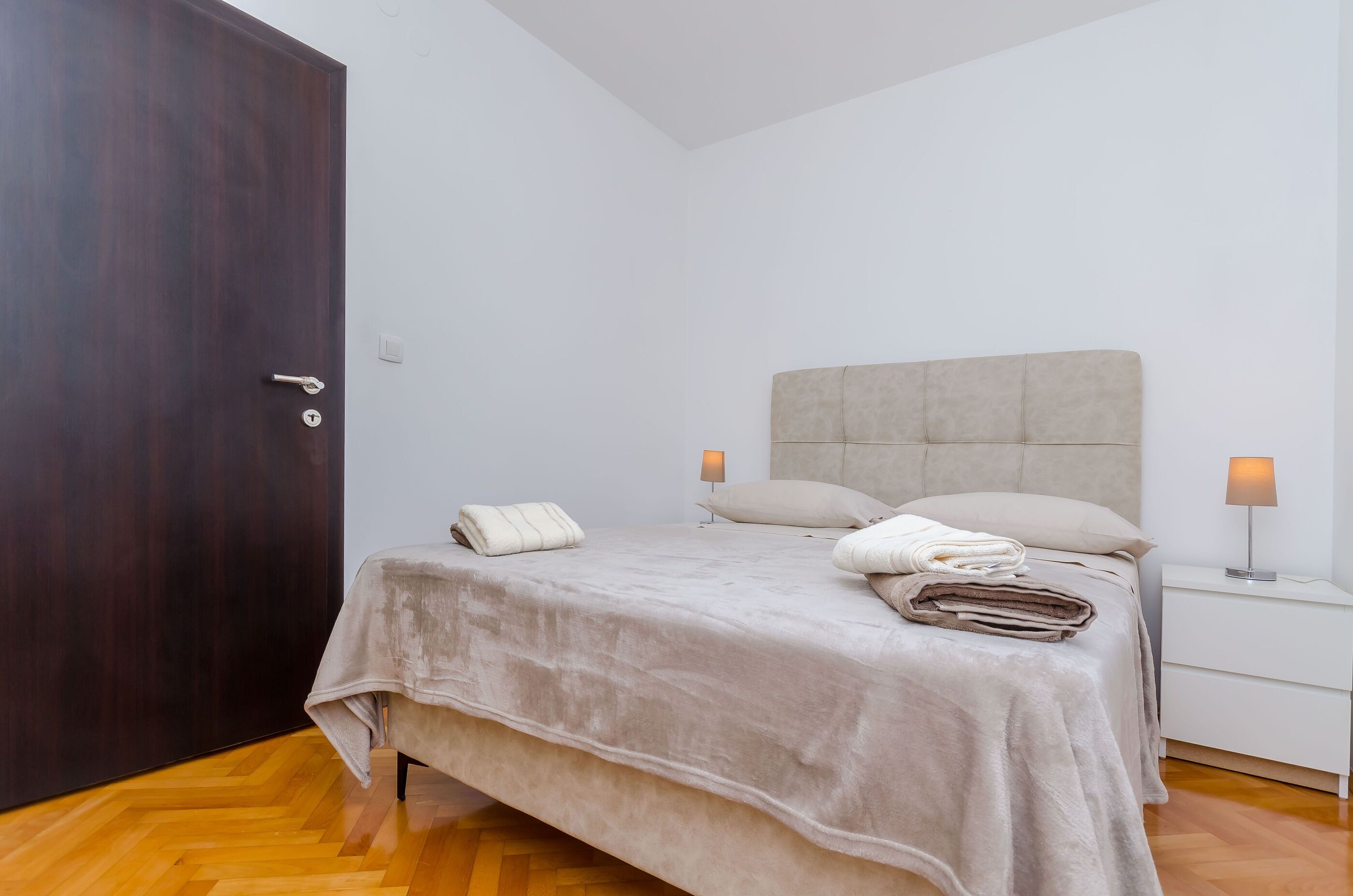 A bedroom with a bed, bedside lamps, a wooden floor, and a dark door.