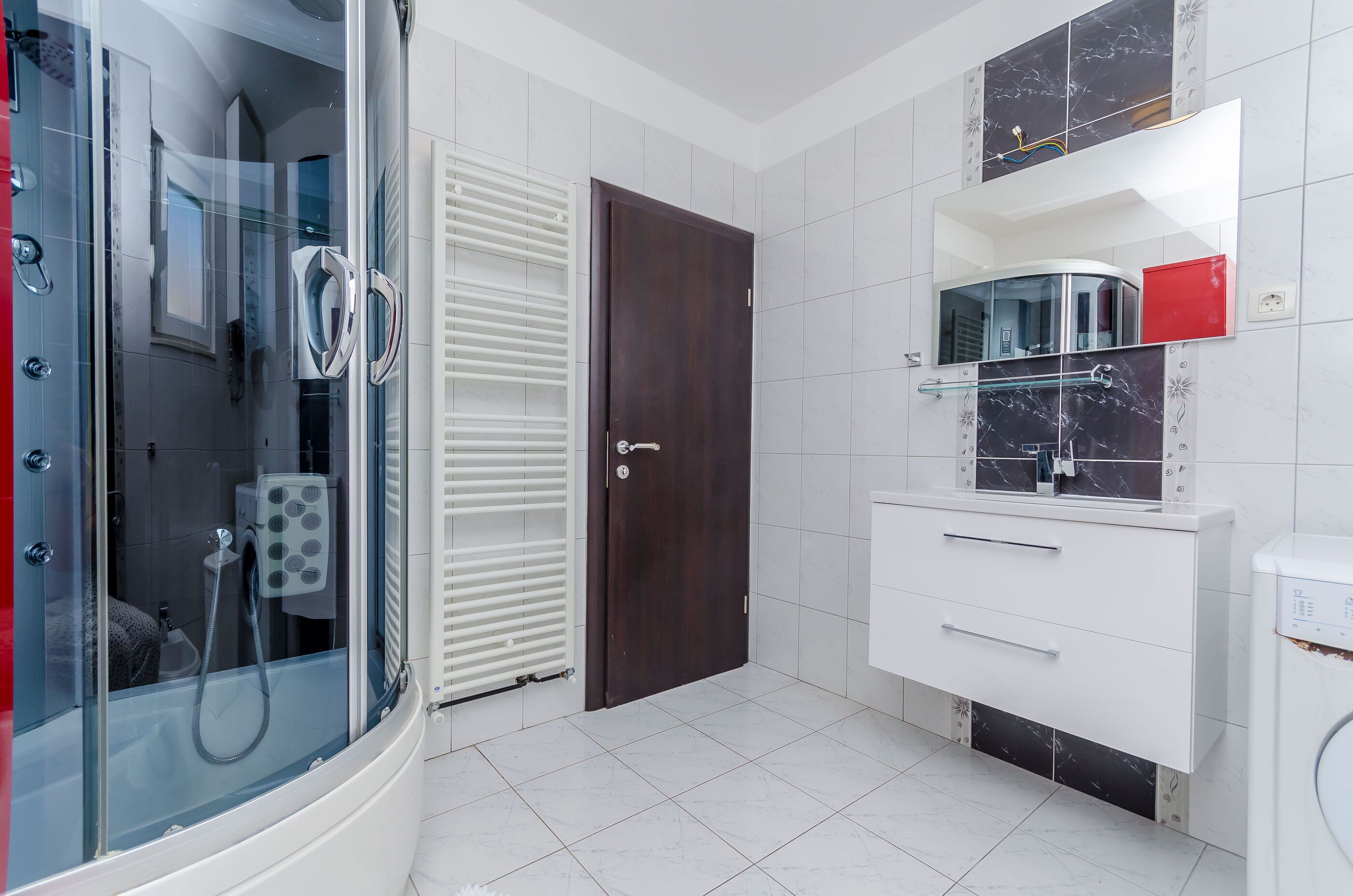 A modern bathroom with a glass-enclosed shower, a dark wooden door, white tiled walls, and a white vanity with a mirror.