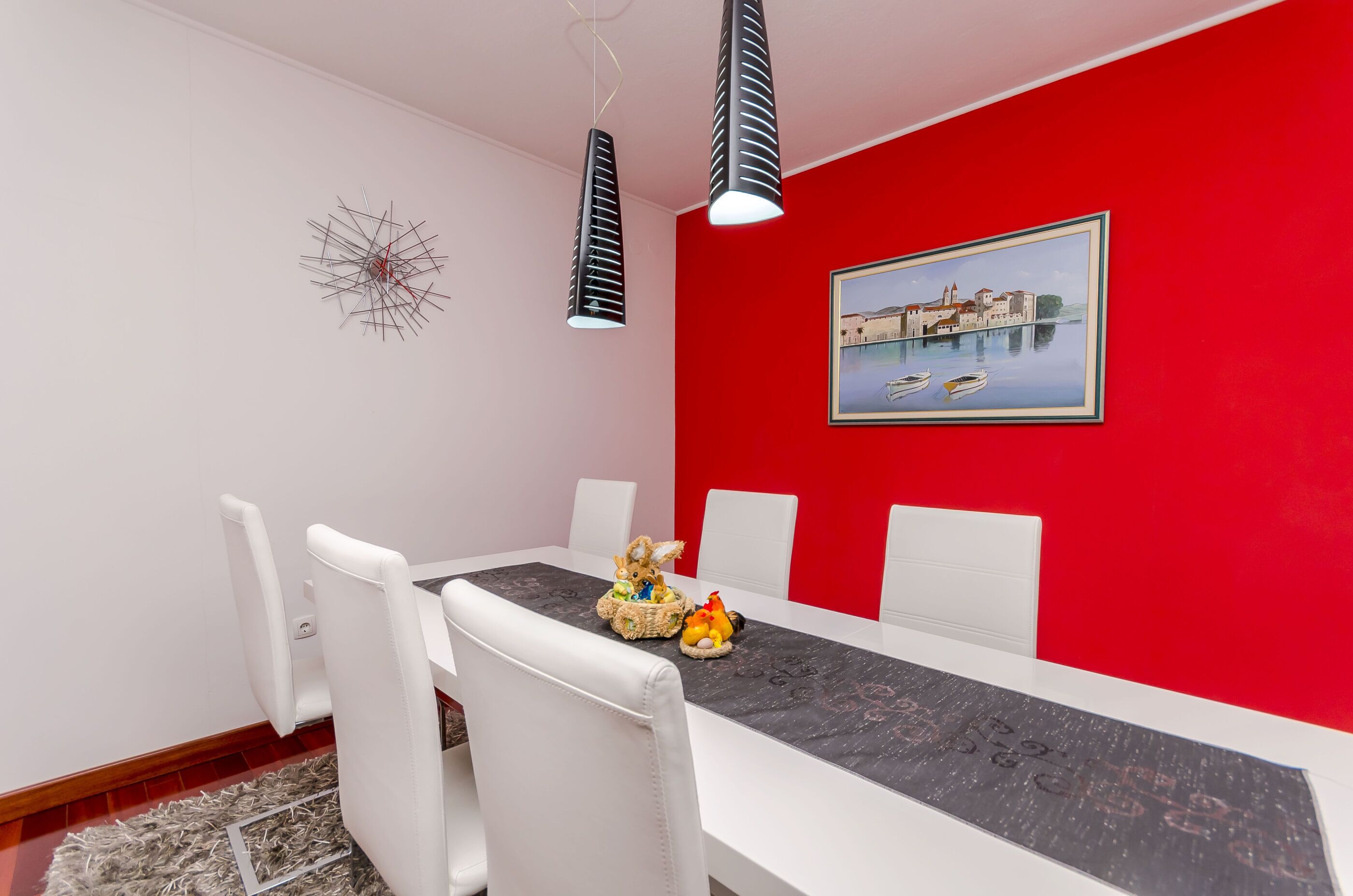 Un coin repas avec un mur rouge, des chaises blanches, une nappe noire et un tableau encadré représentant un paysage de canal.