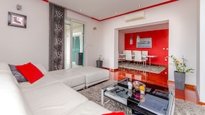 Un salon moderne avec un canapé blanc, une table basse en verre et un coussin décoratif rouge. Un mur d’accent rouge et un petit coin repas avec une table et des chaises blanches complètent l’ensemble.