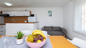 A modern kitchen with white cabinets, a microwave, and a small potted plant. A dining table with a pink bowl of fruit and a wine glass is in the foreground. - Apartments Milena (Lopar)