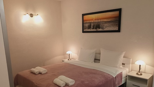 En snyggt bäddad säng med ett rosa överkast, två vita kuddar och två hoprullade handdukar. På väggen ovanför sängen hänger en inramad bild av en solnedgång. Två sänglampor är tända, och en liten låda syns på sängens högra sida.