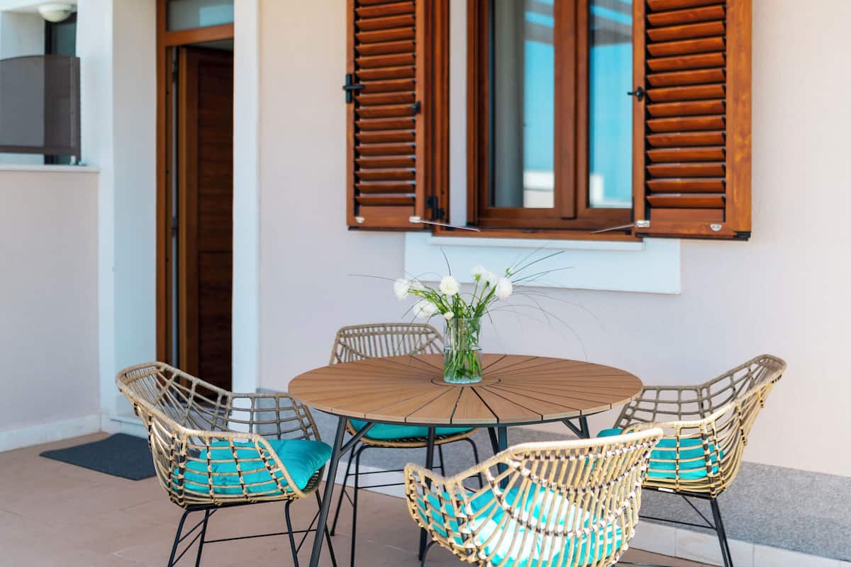 A round wooden table with four wicker chairs, each with teal cushions, and a vase of white flowers on the table.