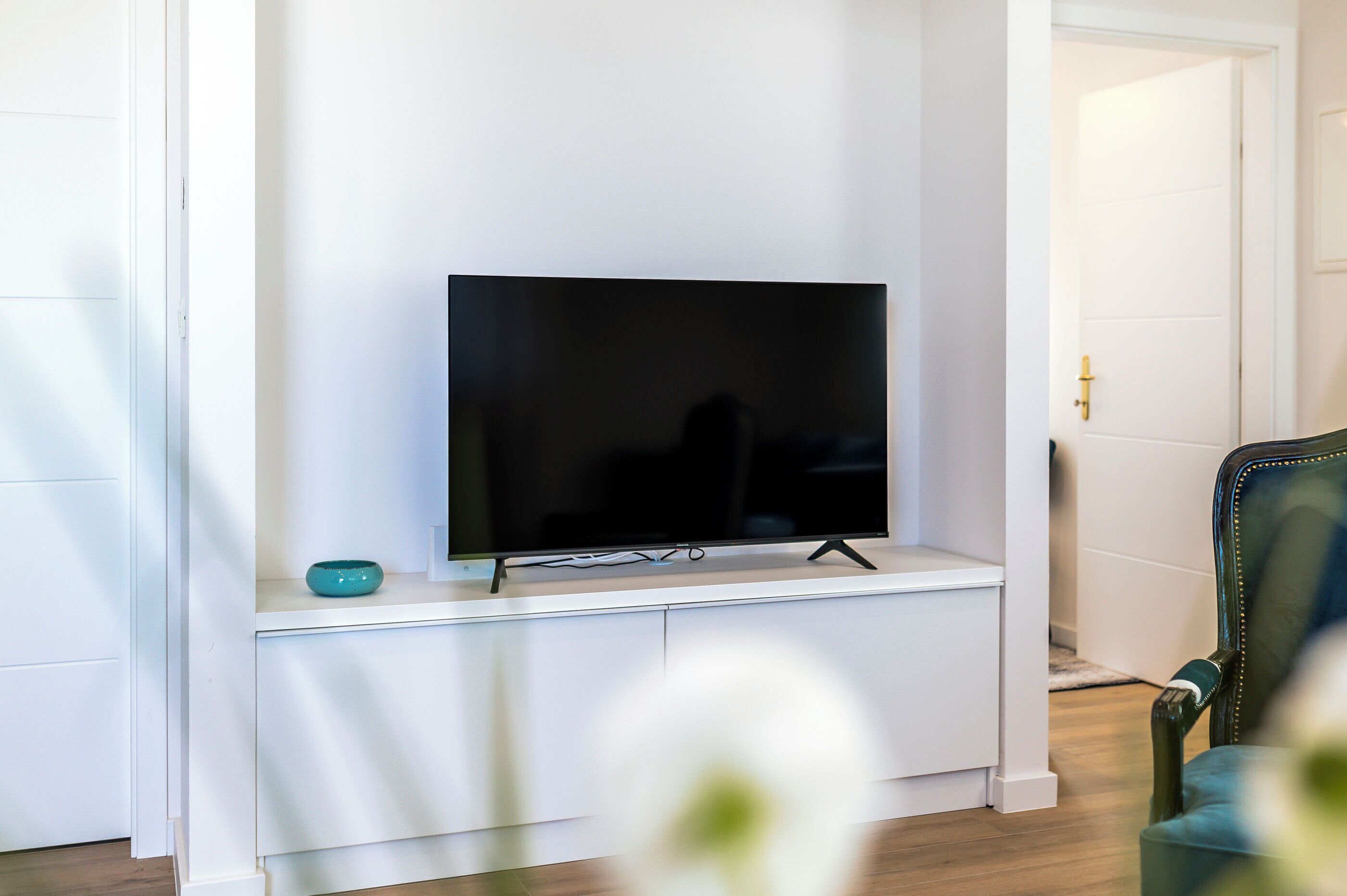 A modern living room with a flat-screen TV on a white stand, a teal bowl on the stand, and a dark green armchair.