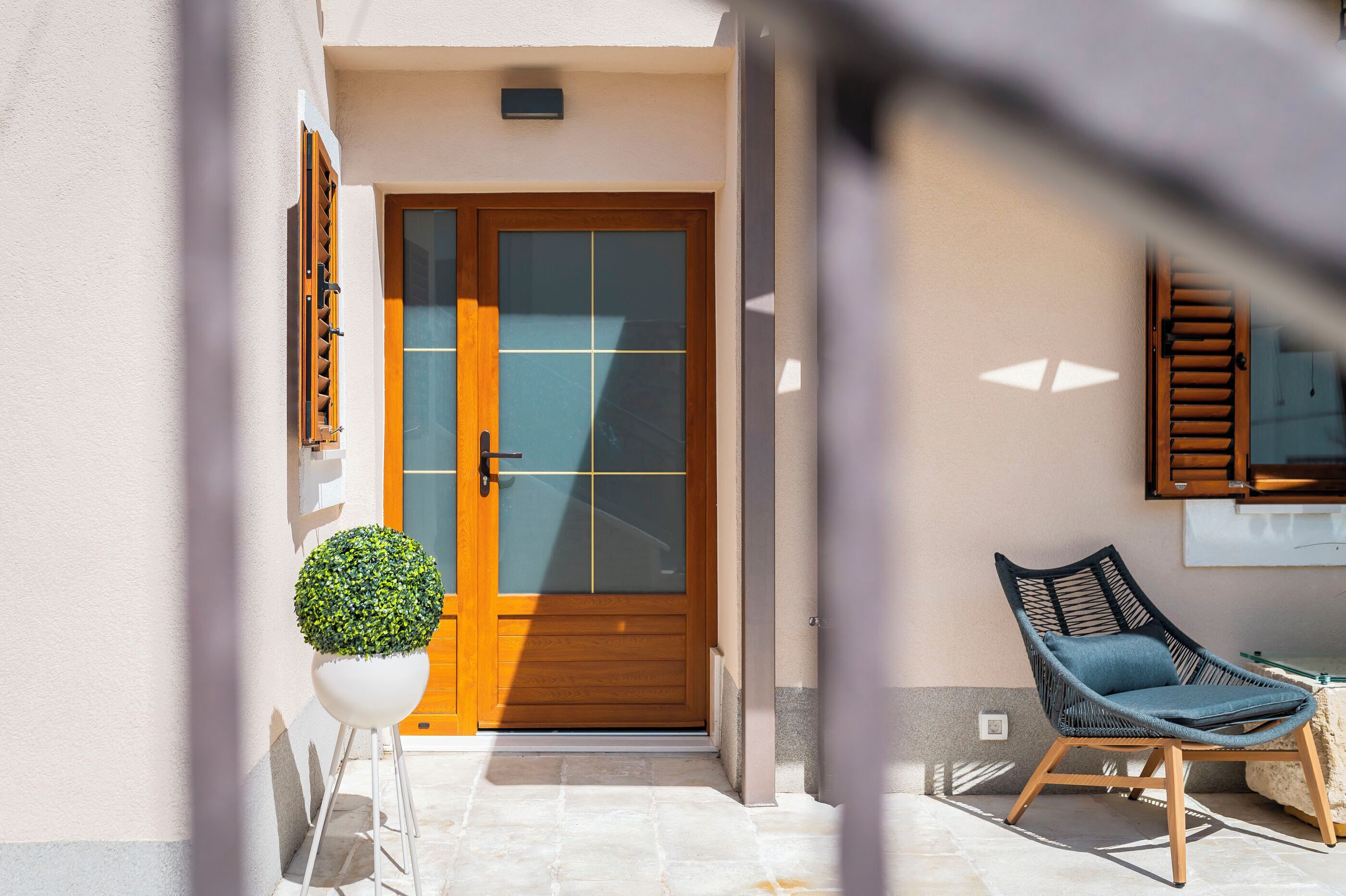 Une porte en bois avec un panneau de verre, une plante en pot et une chaise en osier sur une terrasse carrelée.