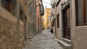 Um estreito beco de calçamento ladeado por edifícios com paredes de pedra e persianas de madeira.