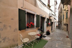 Una calle estrecha con un edificio que tiene una pequeña zona de estar al aire libre con sillas de metal y una mesa, un ventanal con contraventanas verdes y macetas con flores rojas.