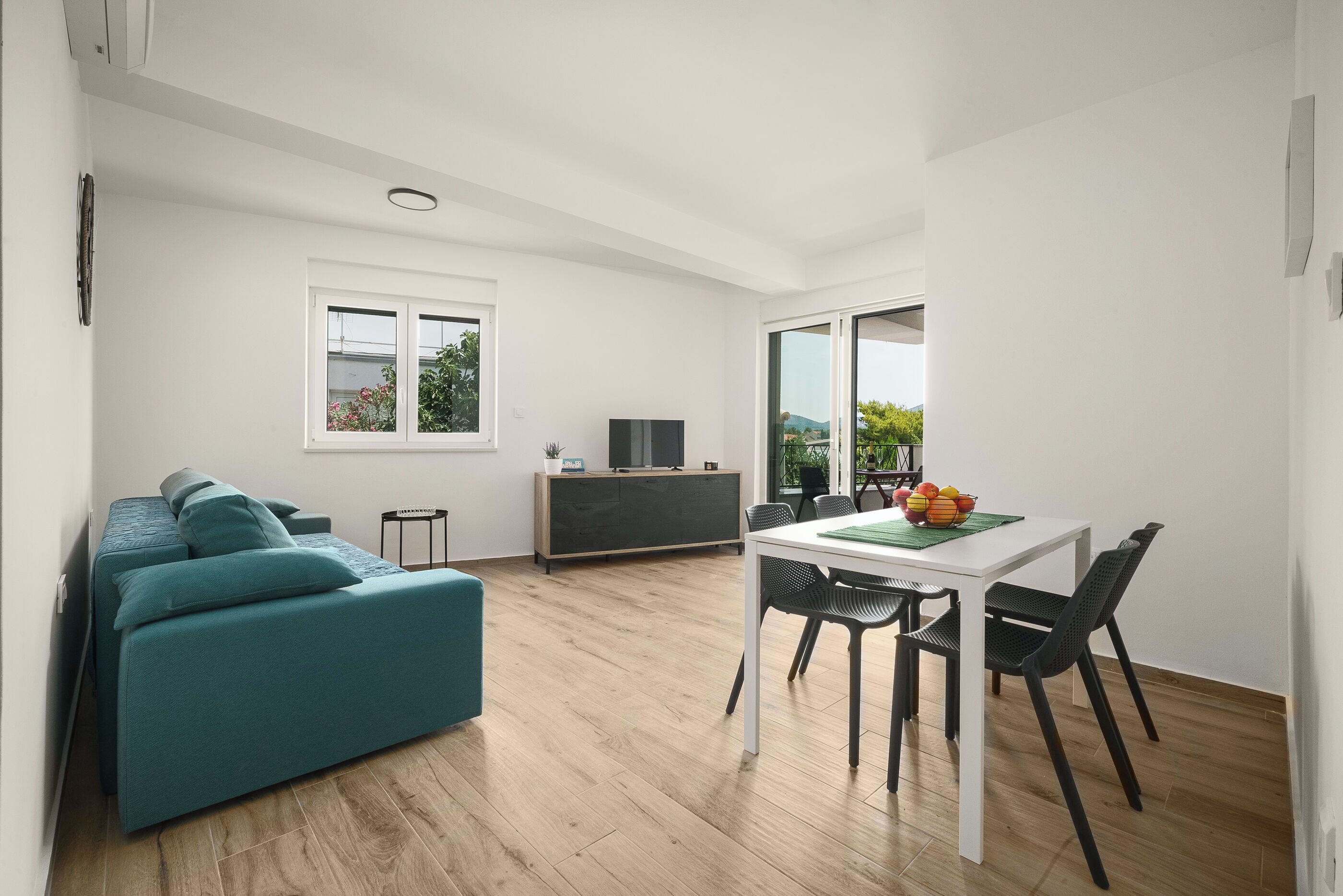 A modern living room with a teal sofa, a dining table with chairs, a TV, and a window with a view of greenery.