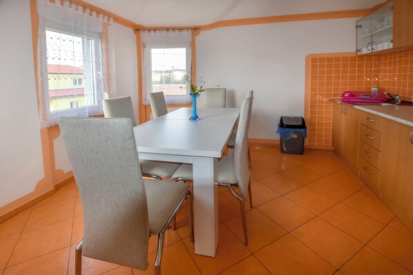 A dining area with a white table, grey chairs, a vase with flowers, and a window with a view of buildings. - House Levanat (Polaca)