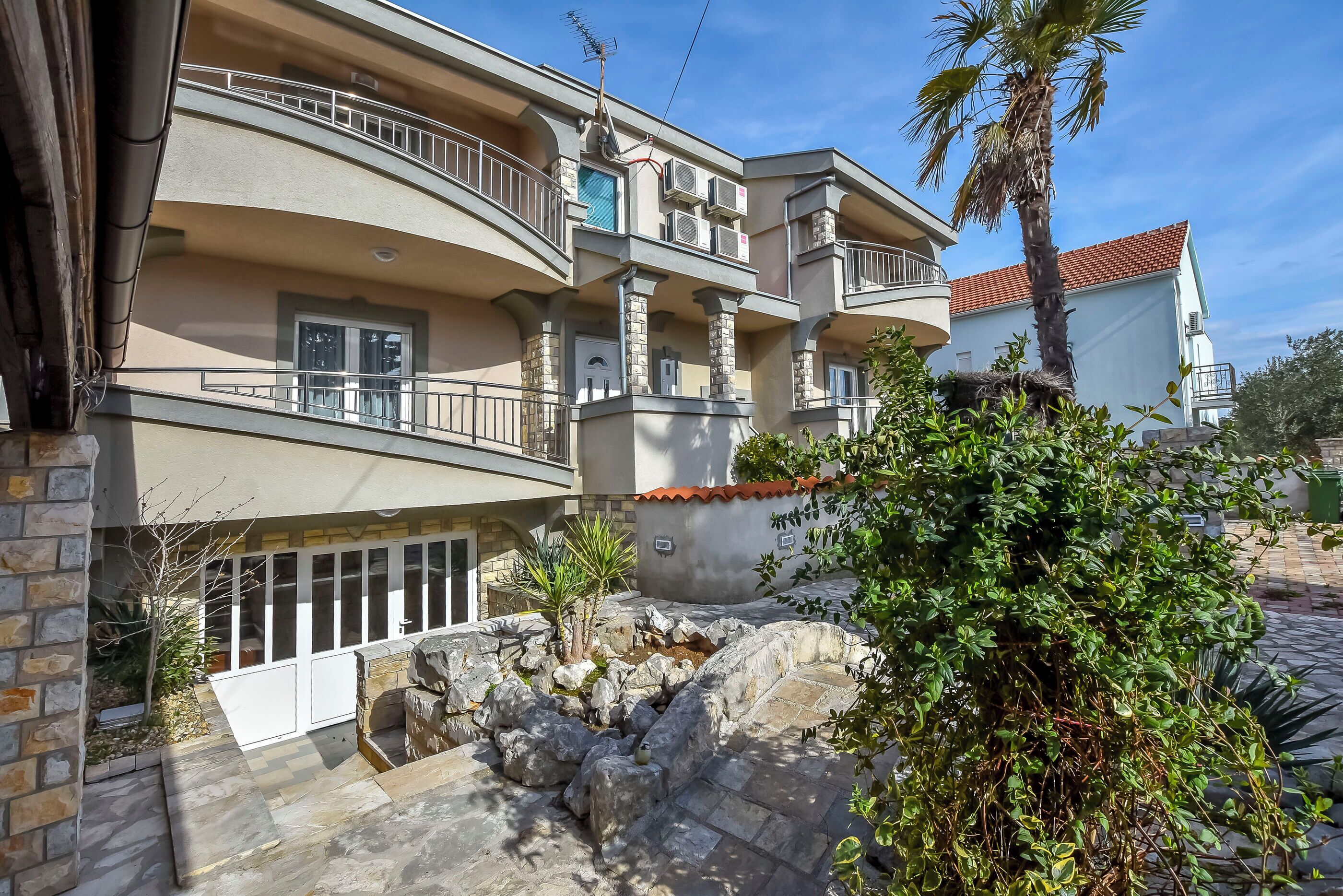 A multi-story residential building with balconies, a stone pathway, and a small garden area.