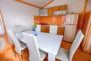 A dining area with a white table and grey chairs, a vase with flowers, and a kitchen with orange tiles and wooden cabinets. - House Levanat (Polaca)