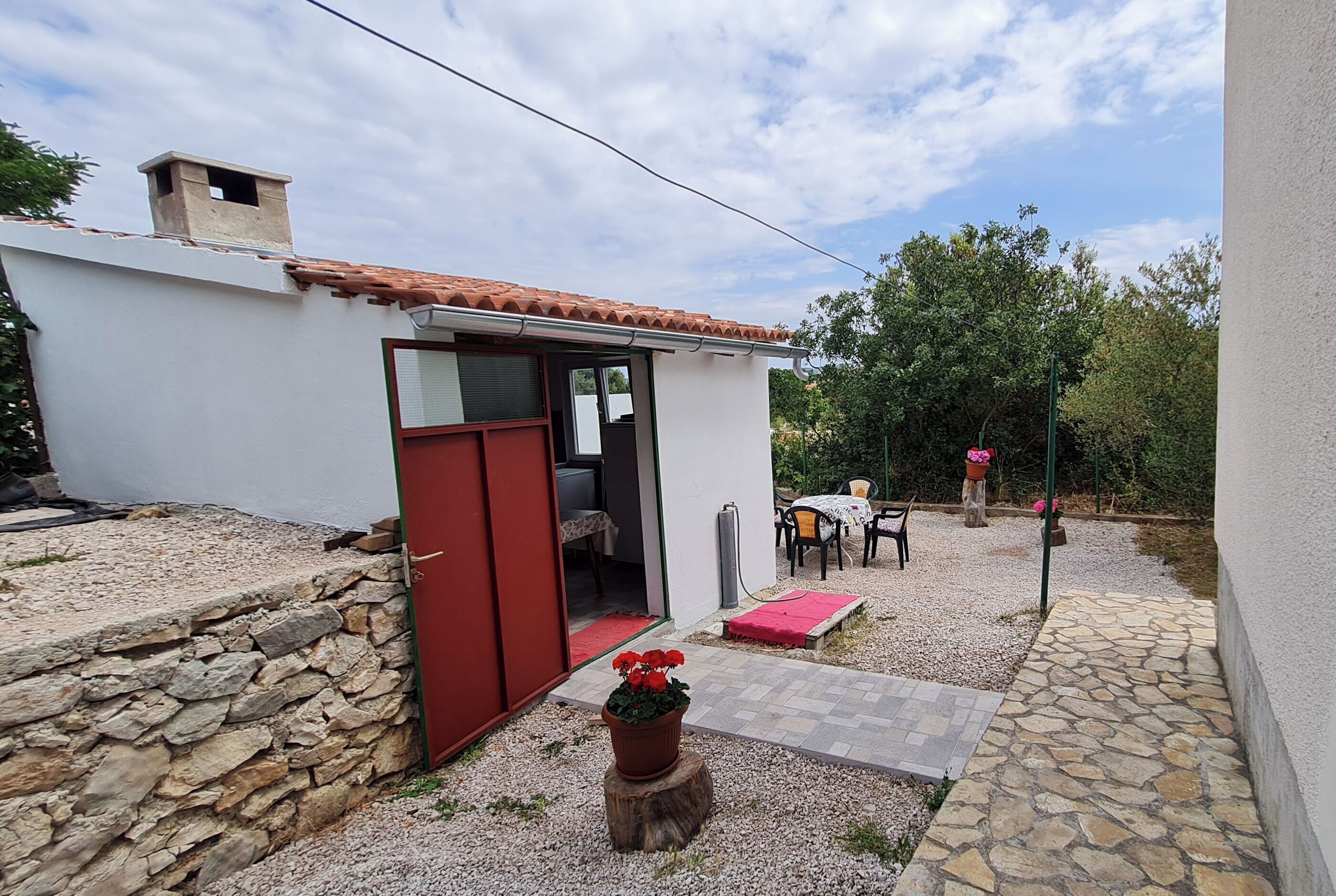 Un pequeño edificio blanco con puerta roja, pared de piedra y jardín con mesa y sillas.
