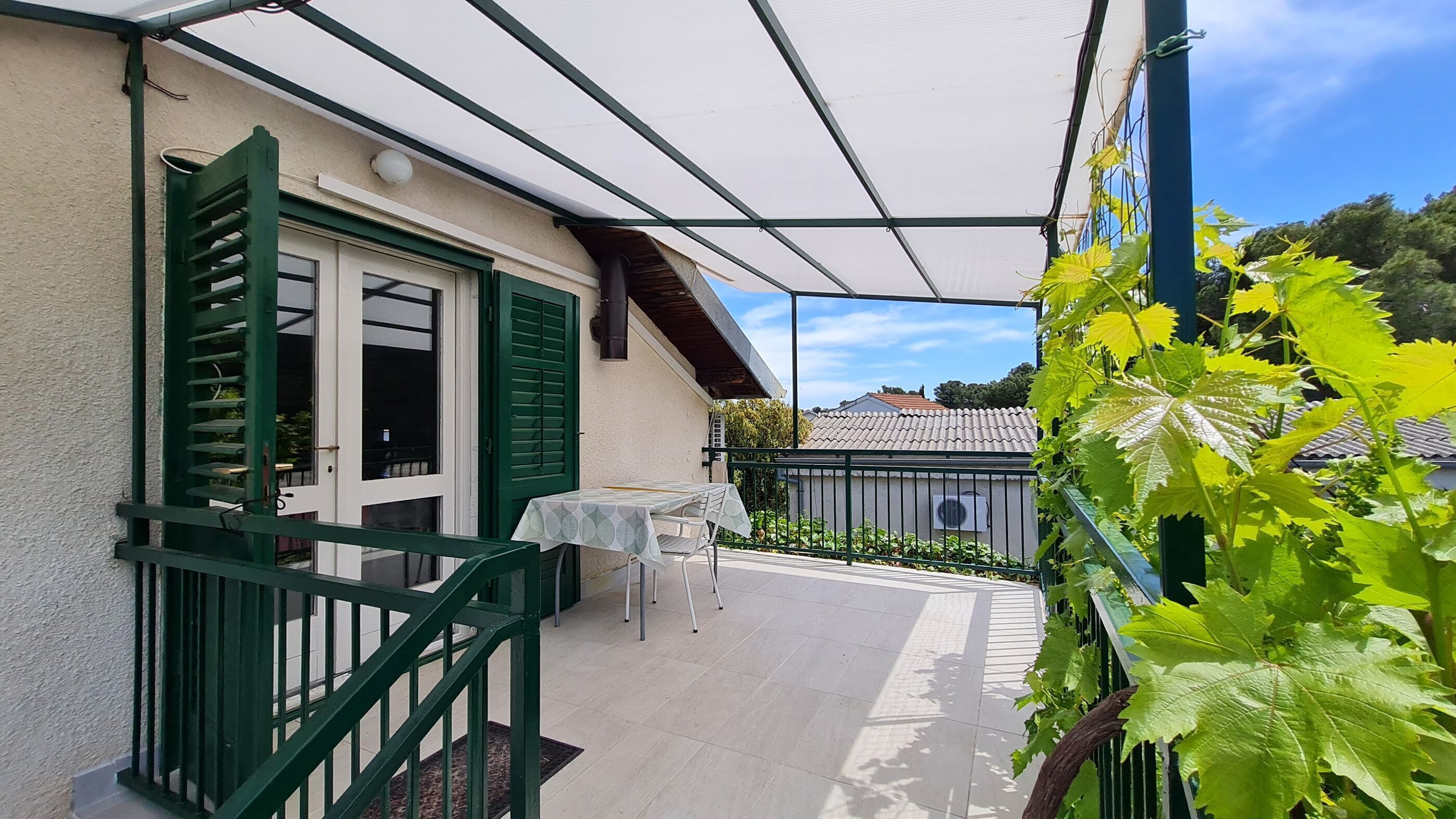 A covered outdoor patio with a table and chairs, a green railing, and a view of the surrounding area.