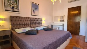 A bedroom with a tufted headboard, a large bed with a purple bedspread, two bedside lamps, a nightstand, a dresser, a mirror, and a wooden door. - Apartment Next to the sea (Jelsa)