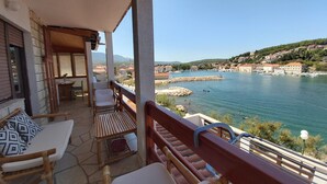 A balcony with wooden furniture, a stone floor, and a view of a coastal town with buildings and a body of water. - Apartment Next to the sea (Jelsa)