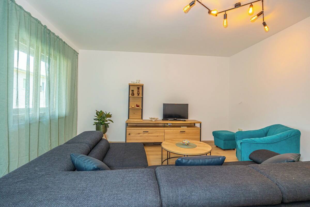 Un salon moderne avec un canapé d’angle, un fauteuil turquoise, une petite table ronde et un téléviseur posé sur un meuble en bois.
